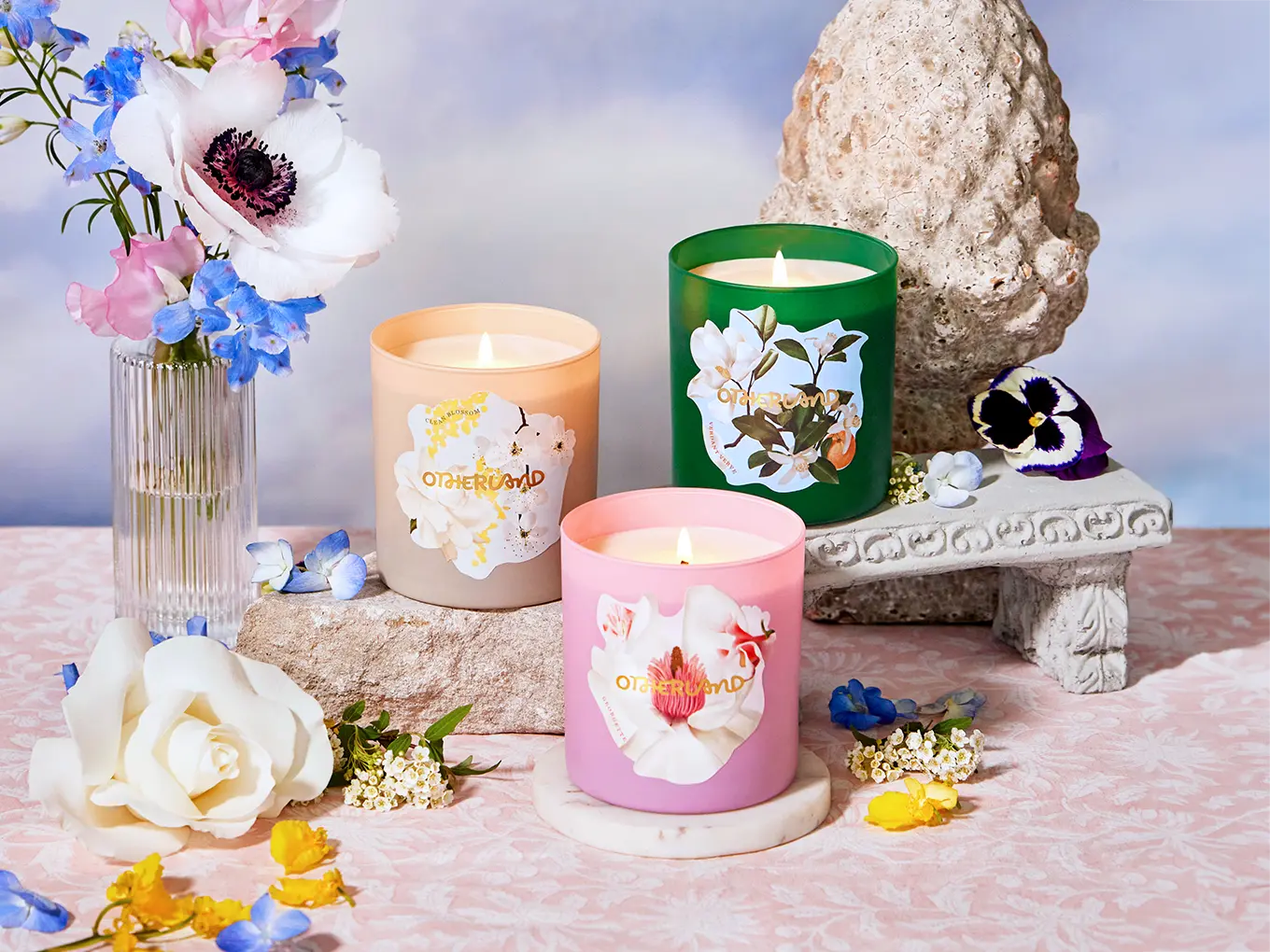 Three scented candles with floral designs on a decorative table with flowers and a stone sculpture.