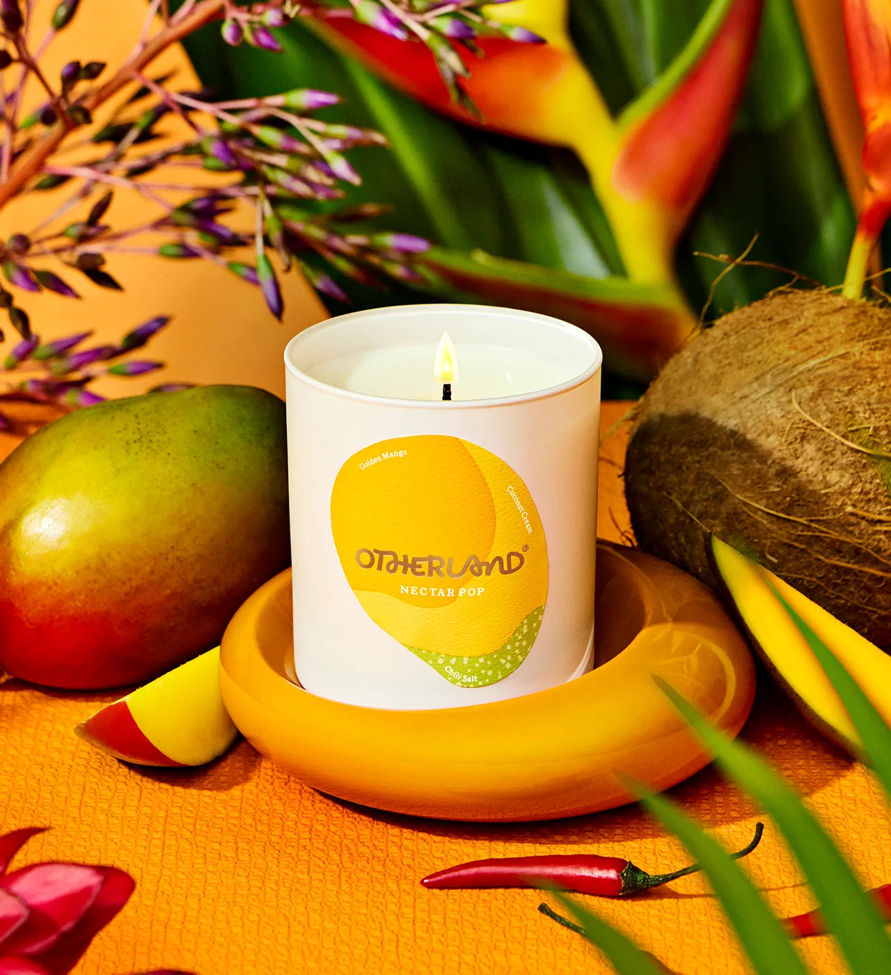 Candle with 'Otherland' branding surrounded by tropical fruits and plants on a warm-toned background - View 2