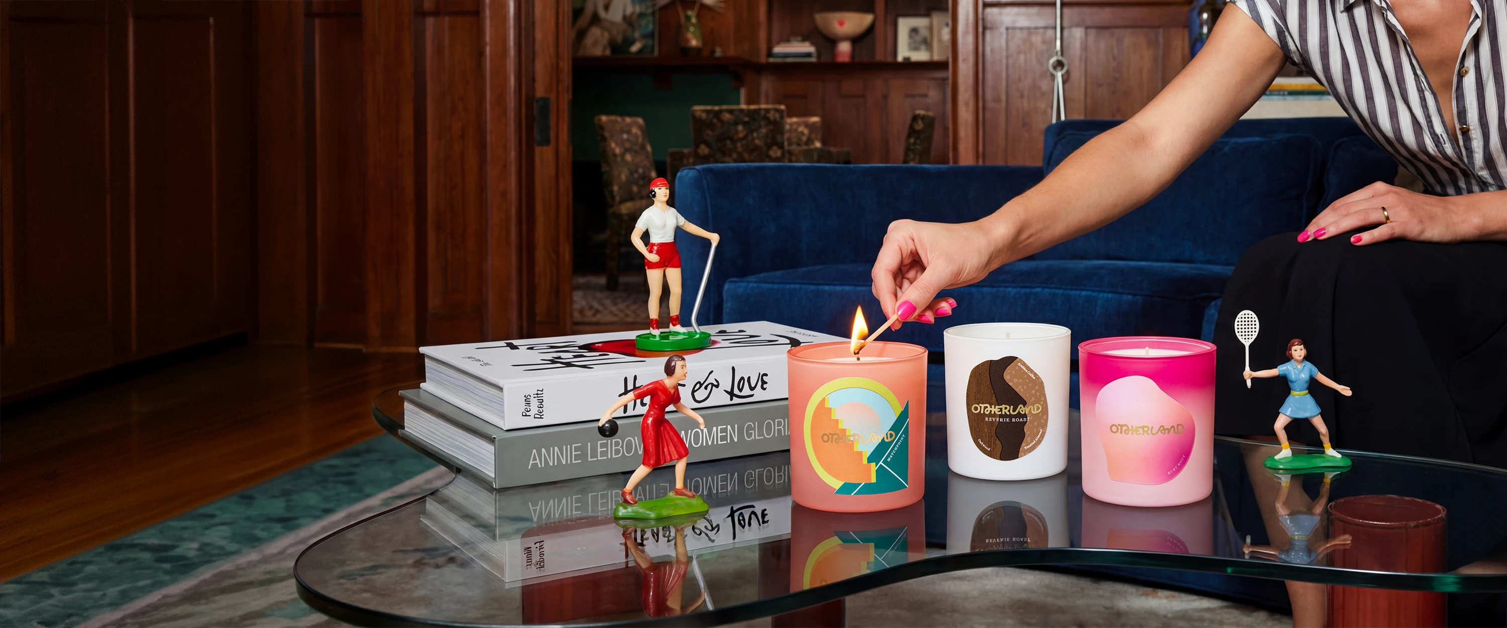 Person lighting a candle on a table with books and decorative items in a cozy living room.