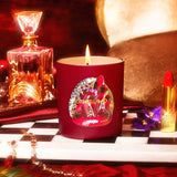 Red candle with a decorative label on a checkered surface, surrounded by a bottle, lipstick, and other items.