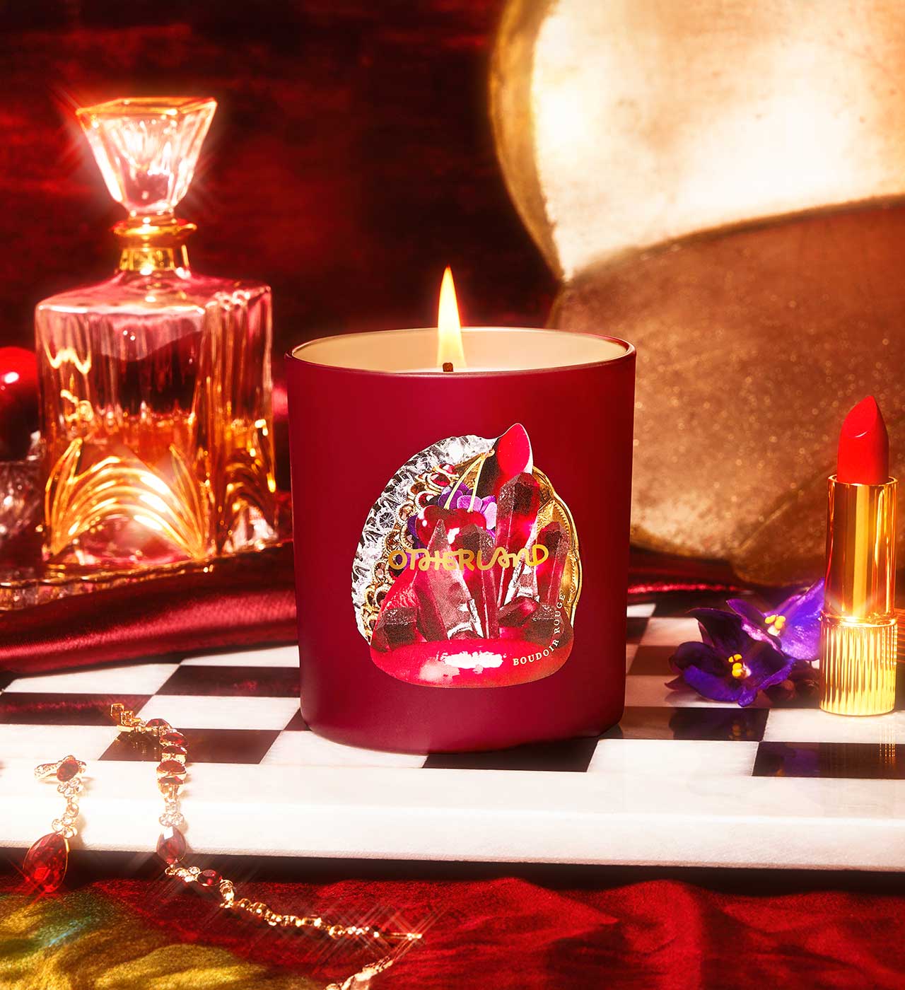 Red candle with a decorative label on a checkered surface, surrounded by a bottle, lipstick, and other items. - View 2