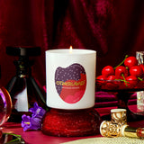 Lit candle with 'Otherland' branding on a red surface with cherries and perfume bottles.