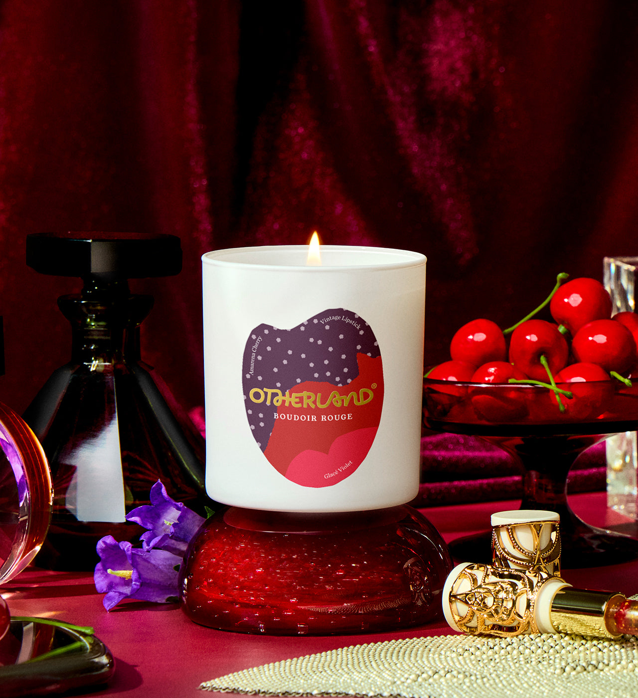 Lit candle with 'Otherland' branding on a red surface with cherries and perfume bottles. - View 2