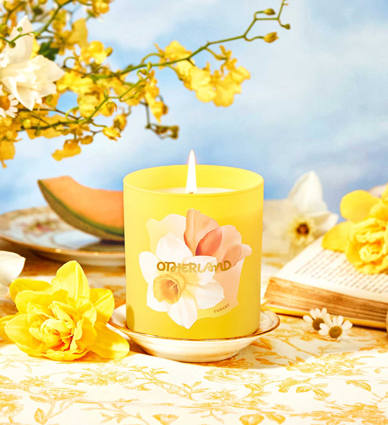 Yellow candle with floral design on a table with yellow flowers and a blue sky background - View 2