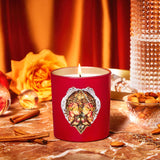 Red candle with decorative lid on a table with cinnamon sticks, rose, and nuts.