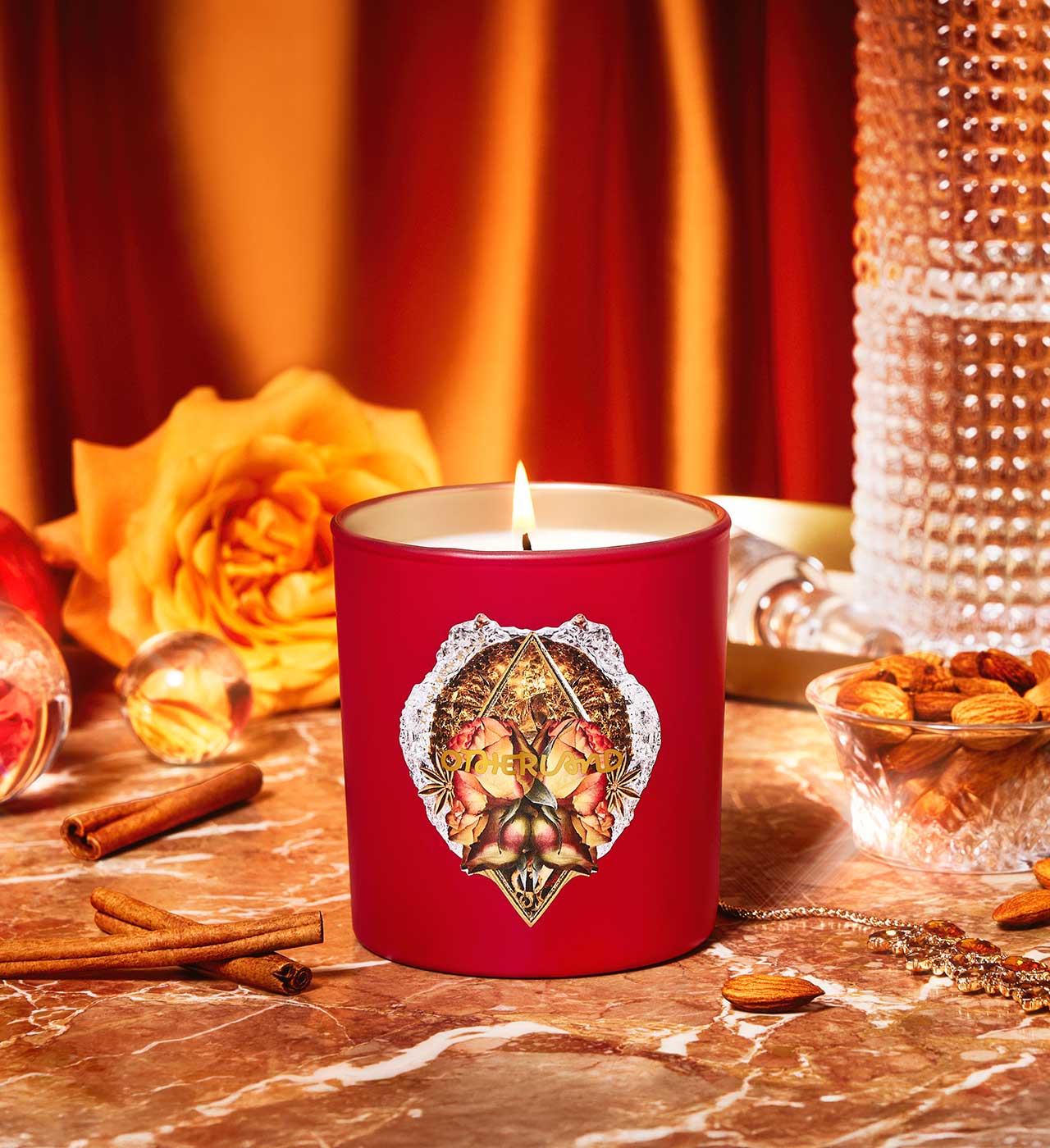 Red candle with decorative lid on a table with cinnamon sticks, rose, and nuts. - View 2