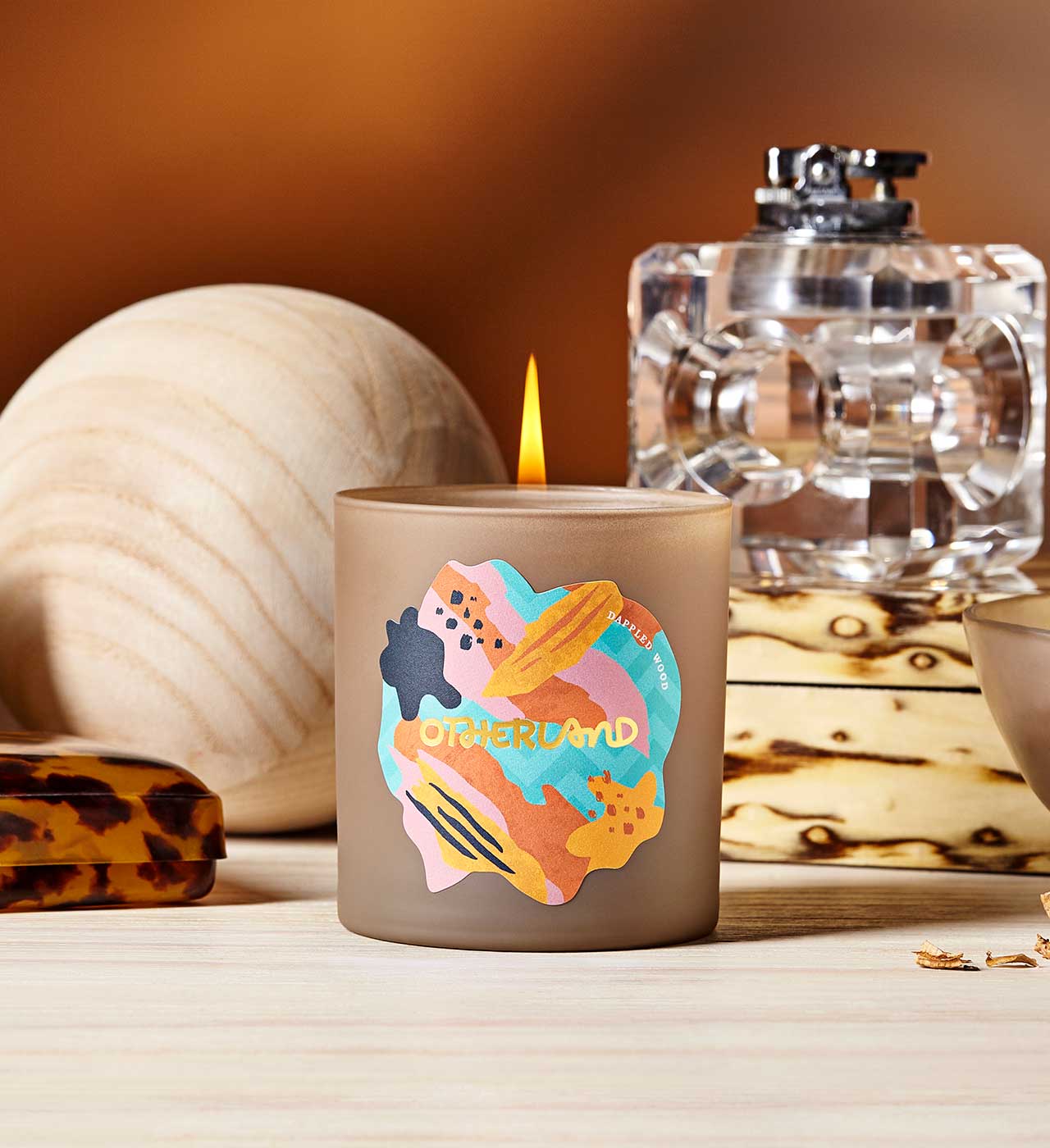 Candle with colorful design and 'Otherland' branding on a wooden surface. - View 2