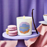 Candle with a dessert design on a purple stand with macarons and a vanilla bean against a purple curtain.