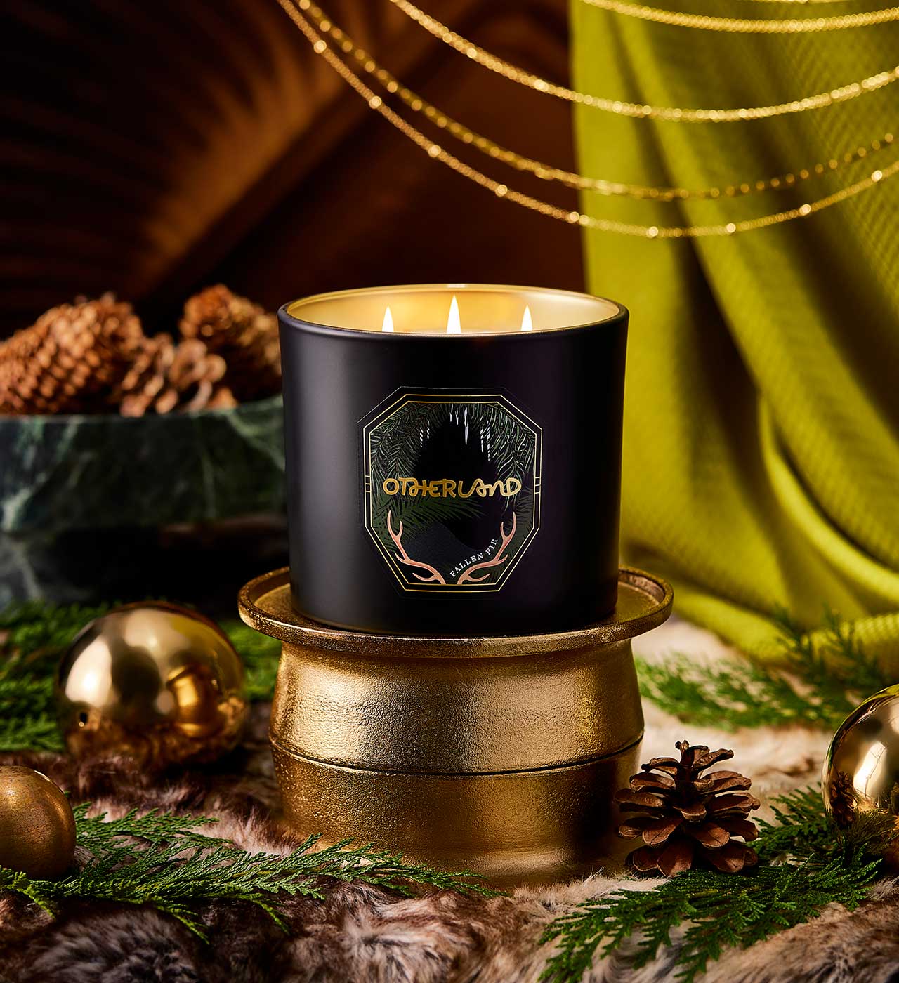 Black candle with 'Otherland' branding on a decorative surface with greenery and gold accents. - View 2