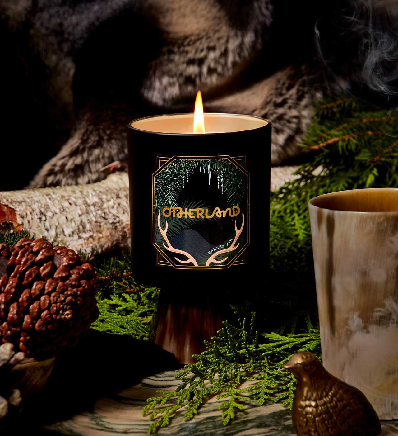 Candle with 'Otherland' branding in a natural setting with greenery and a log. - View 2