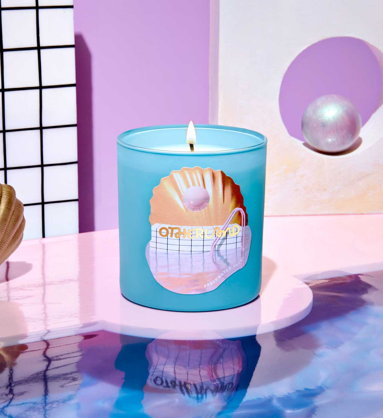 Blue candle with a shell design on a reflective surface with a pink and white background - View 2