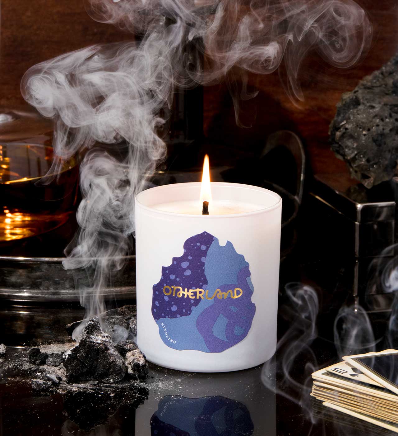 Candle with 'Otherland' logo on a dark background with smoke and rocks. - View 2