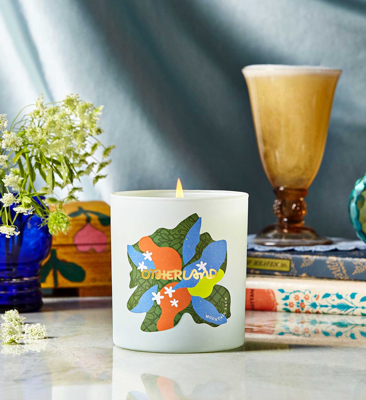 Candle with colorful design on a table with books and flowers - View 2