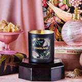 Black candle with gold lid on a decorative stand, surrounded by a pink lamp, books, and a colorful floral pattern.