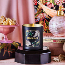 Black candle with gold lid on a decorative stand, surrounded by a pink lamp, books, and a colorful floral pattern.