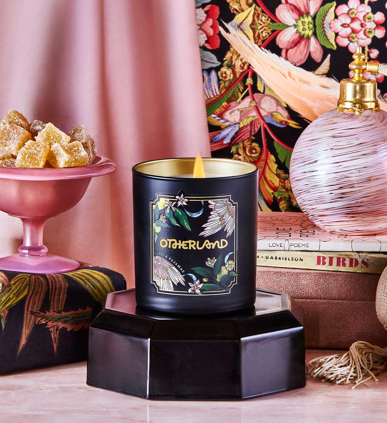 Black candle with gold lid on a decorative stand, surrounded by a pink lamp, books, and a colorful floral pattern. - View 2